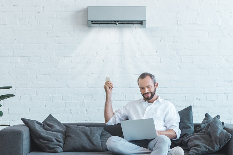 Video - 4 Amazing Benefits of Ductless Units. Man sitting on couch with laptop on lap. ductless air conditioning unit on wall.|