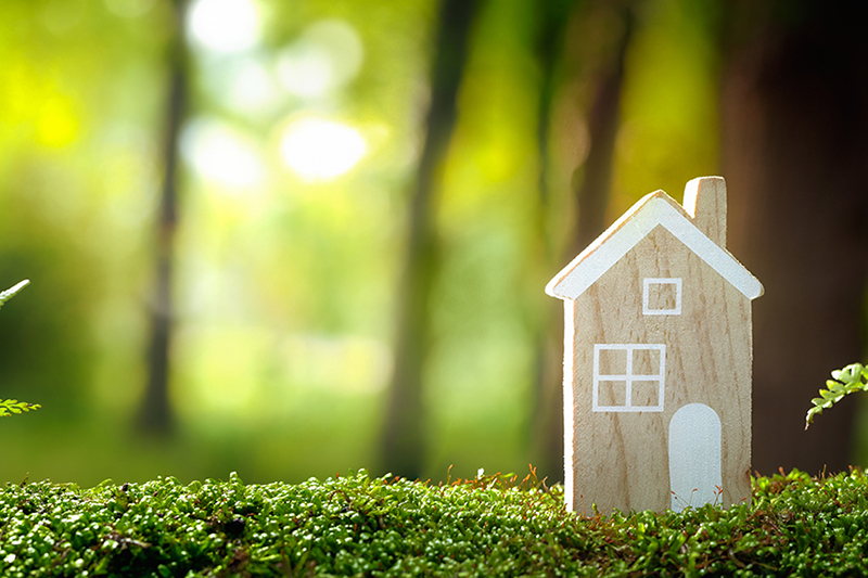 Top 5 Benefits of Installing a Geothermal System. Photo of a wooden toy home placed on artificial grass with a blurry background of the woods.